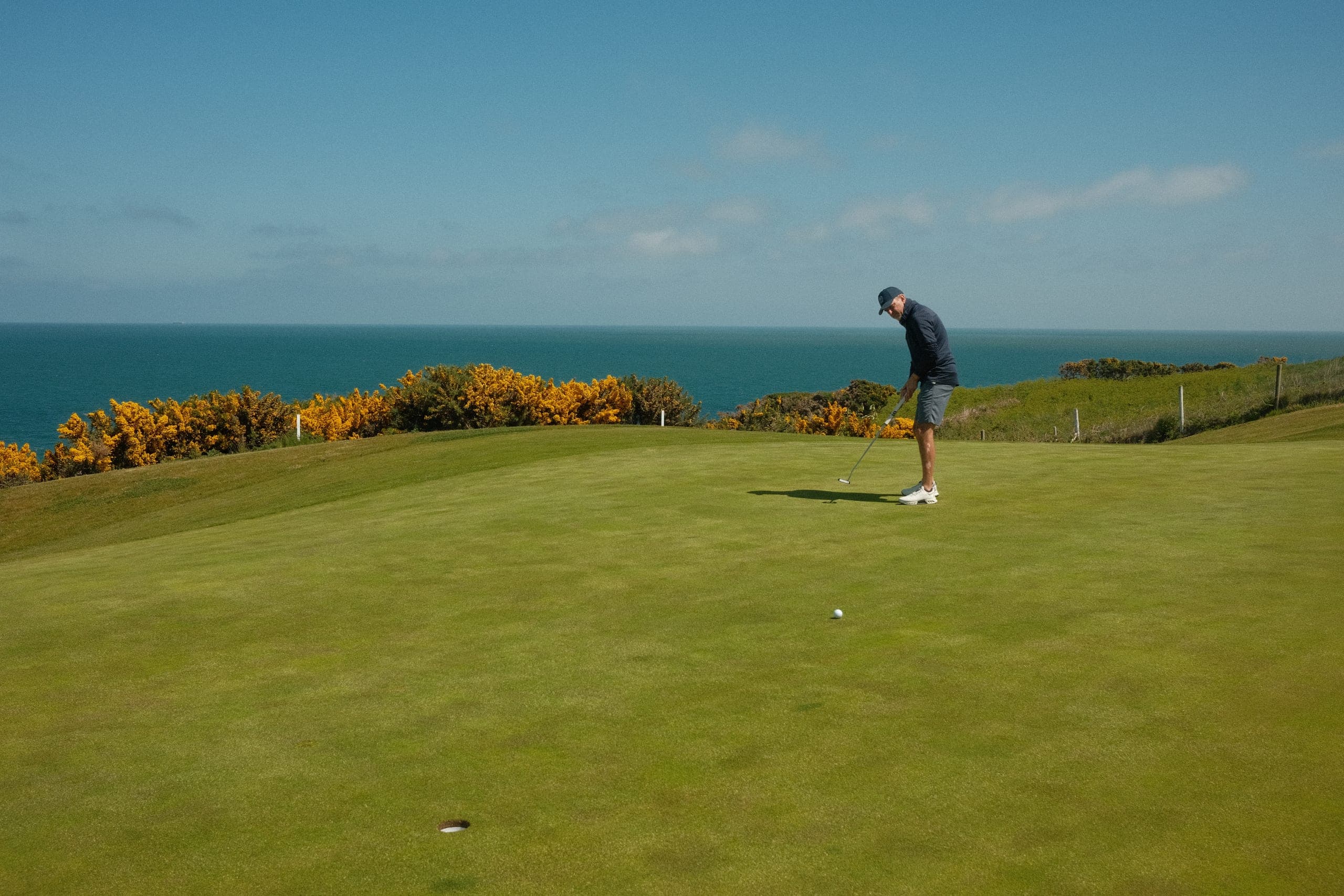 A player putting on a links green, yellow gorse in bloom and the sea beyond.