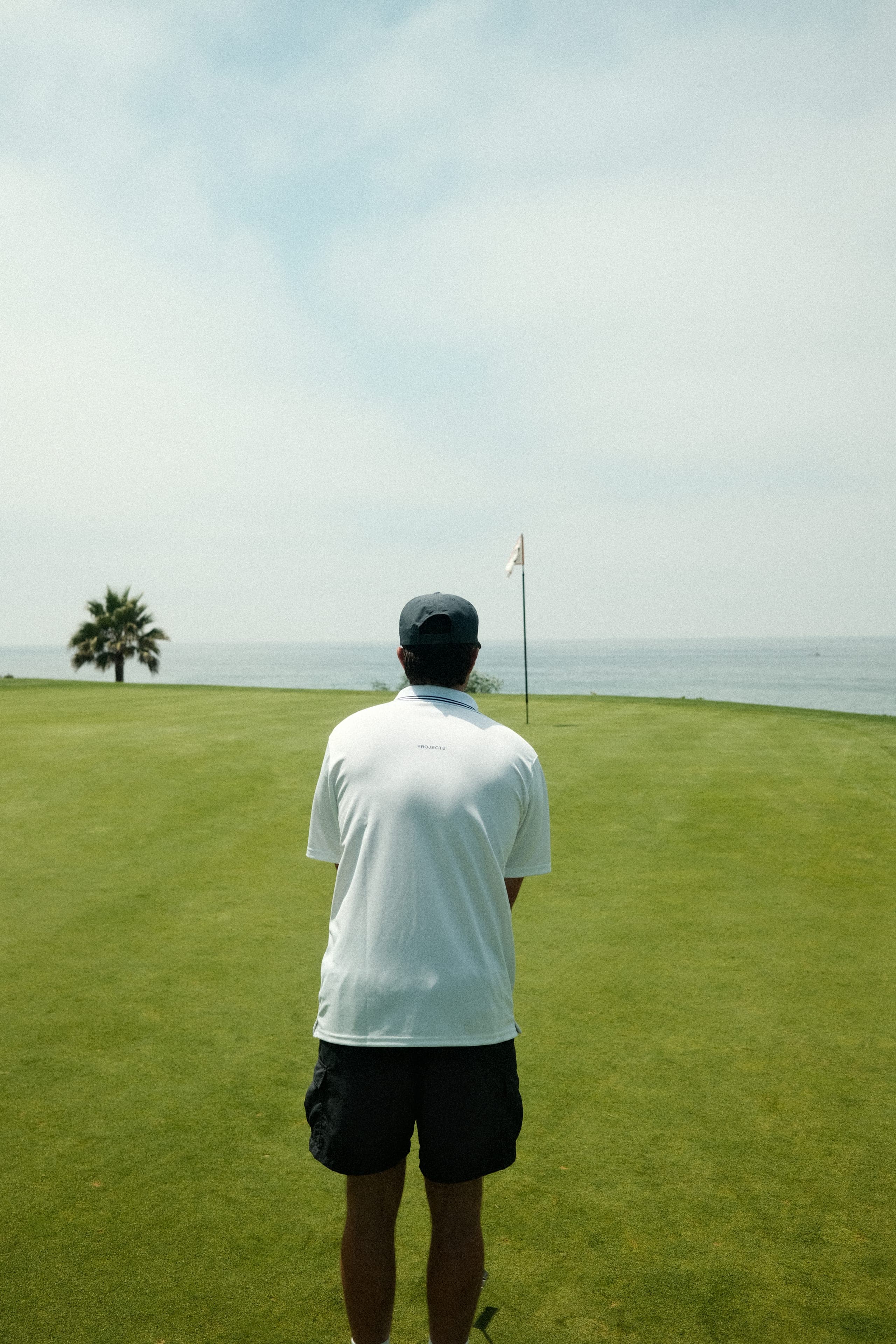 A player seen from behind in a white polo, facing the flag and the sea.