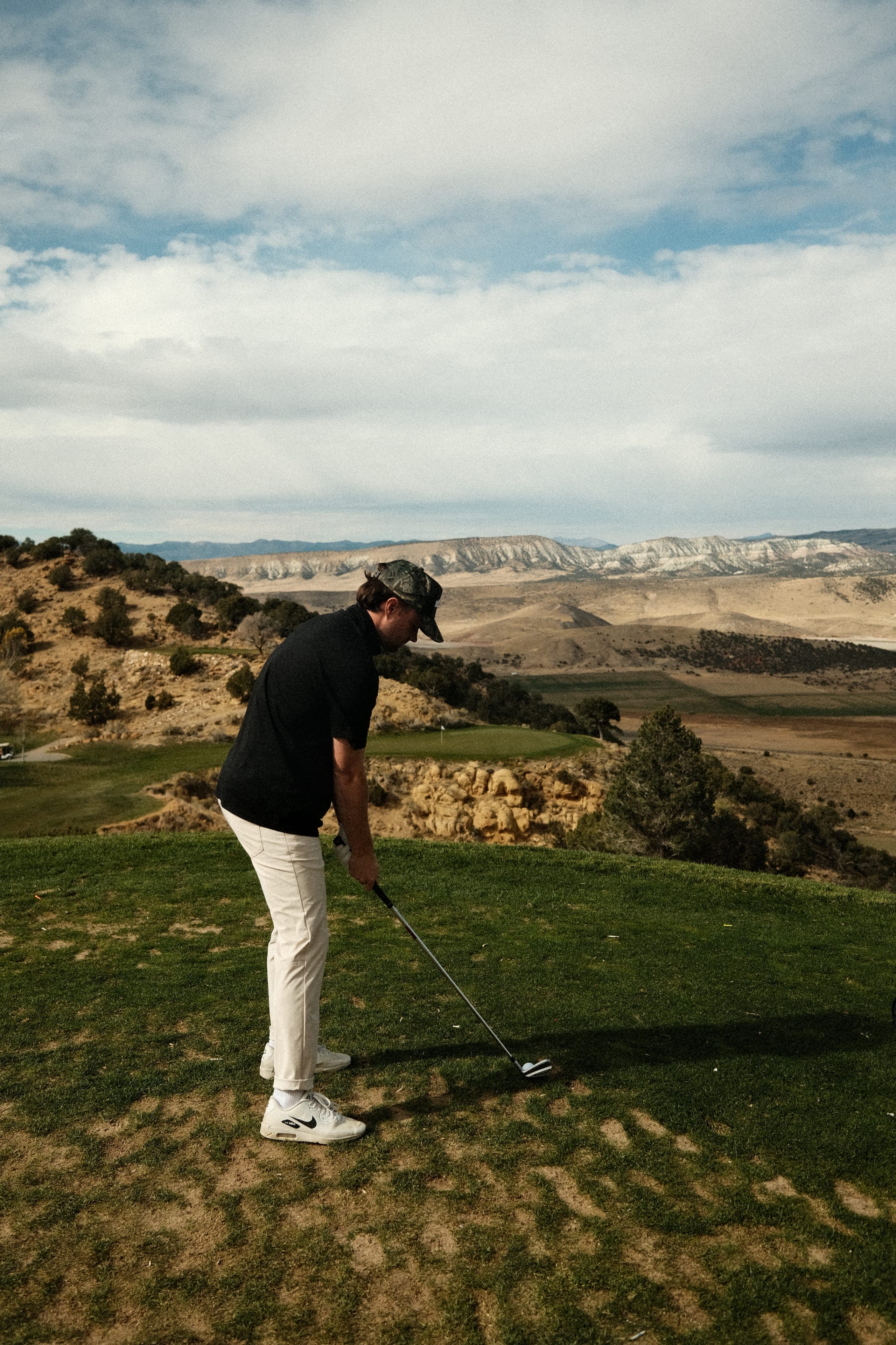 A tee shot in a black polo and stone trousers, desert mountains behind.