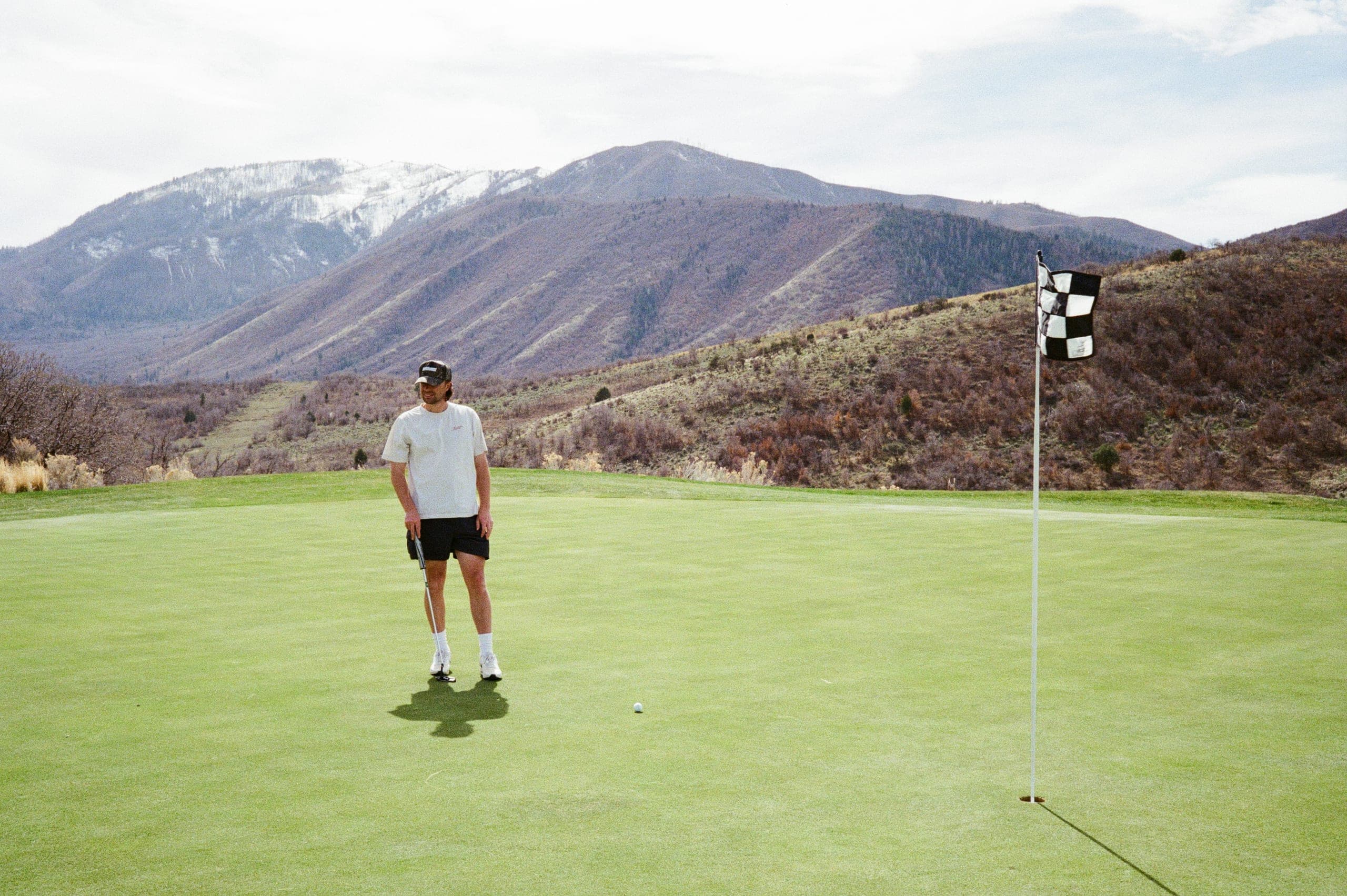 A player on a green beneath snowed mountains, checkered flag flying.