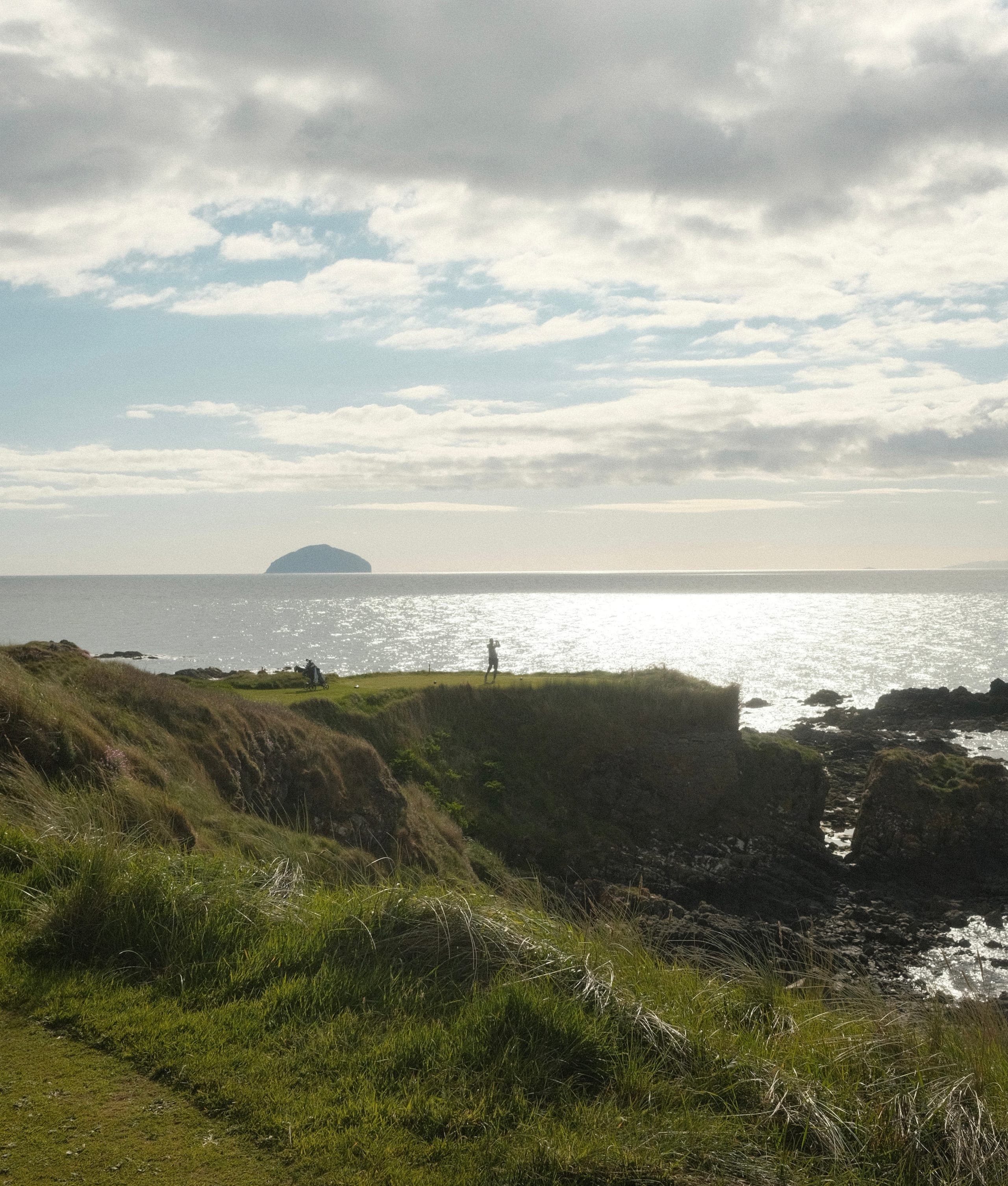 A player teeing off from a clifftop on the Ayrshire coast, Ailsa Craig rising from the sea beyond.