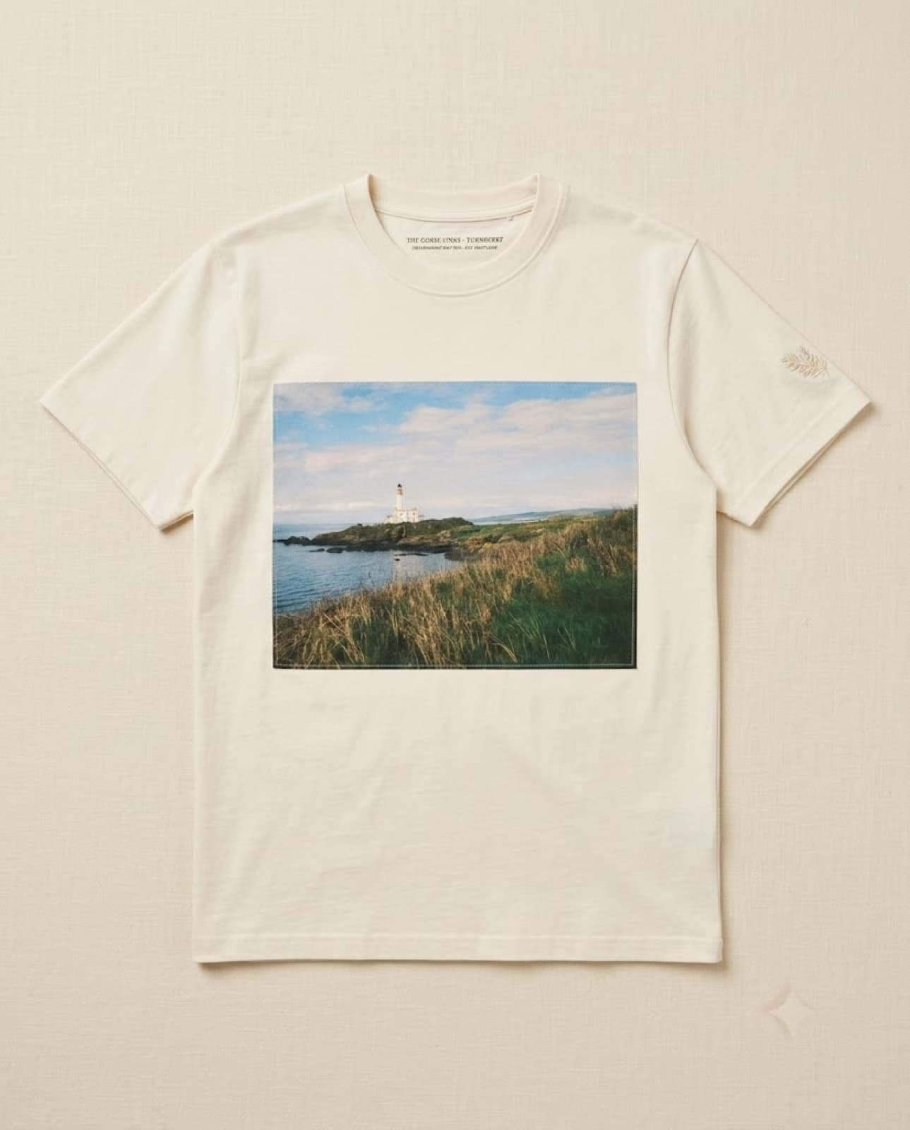 A heavyweight cream tee with a muted duotone coastal links photograph printed at the chest, laid flat on cream linen.