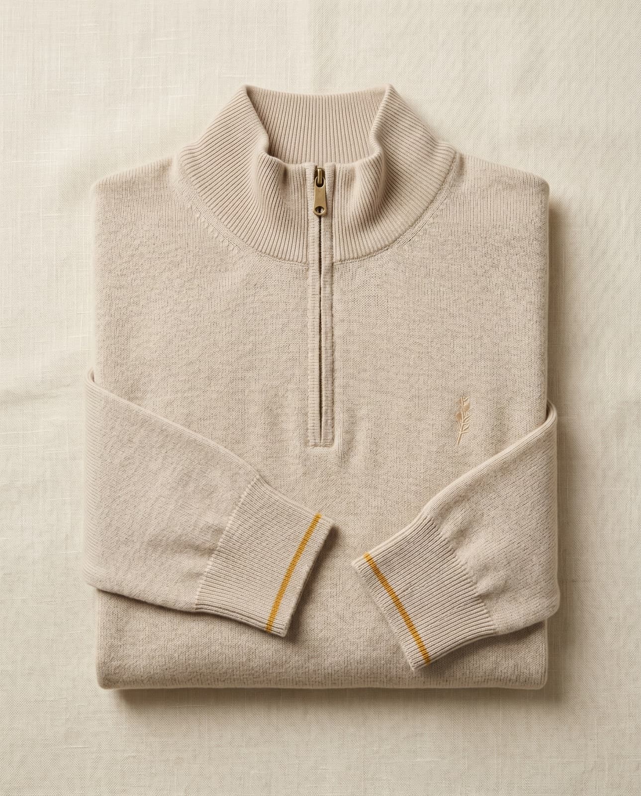 A solid oatmeal merino-cashmere quarter-zip with a raw brass pull, folded on cream linen.